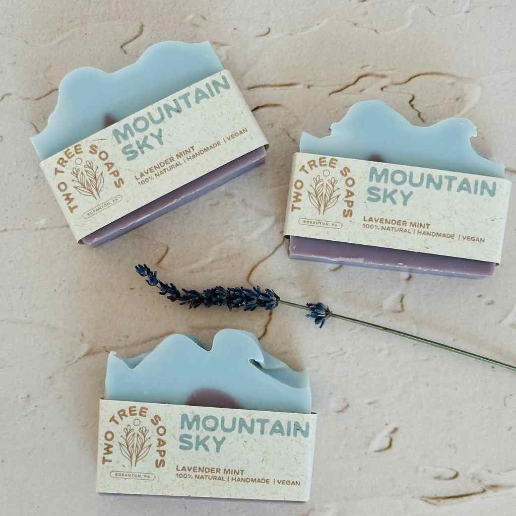 Three bars of 'Two Tree Soaps Mountain Sky' soap in their packaging on a textured surface with a lavender sprig.