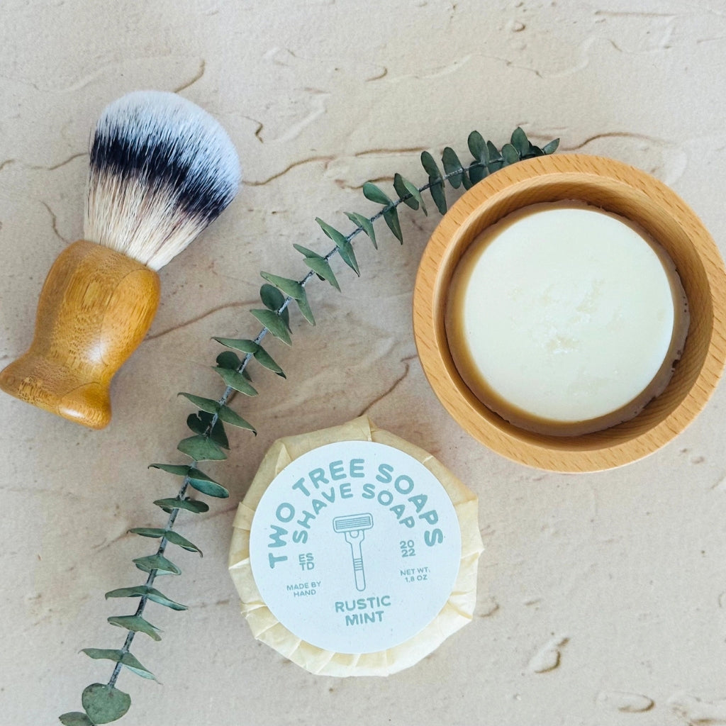 Two Tree Soaps Rustic Mint shave soap bar shown with a wooden-handled shaving brush and a small wooden bowl on a light stone surface, accented by a green eucalyptus sprig.