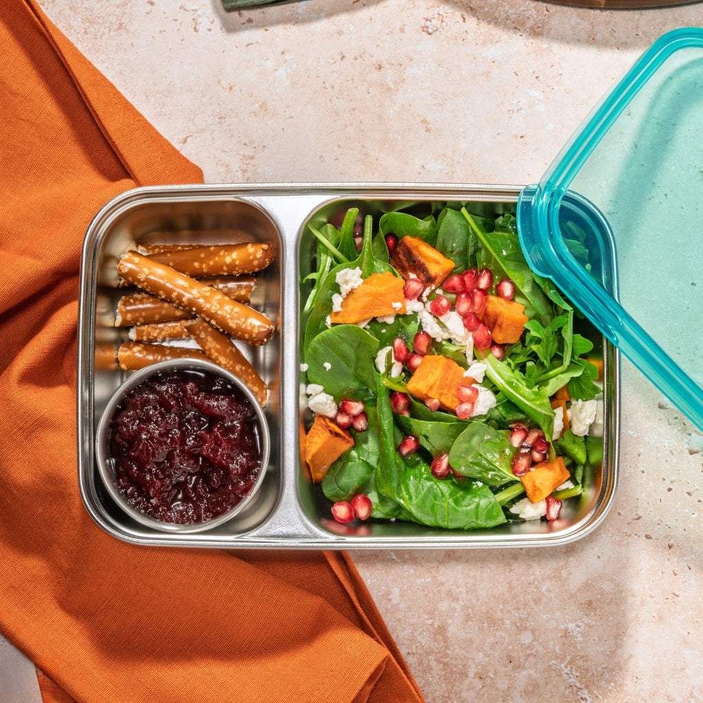 Metal lunch container with salad, pretzels, and a side of sauce on an orange napkin.