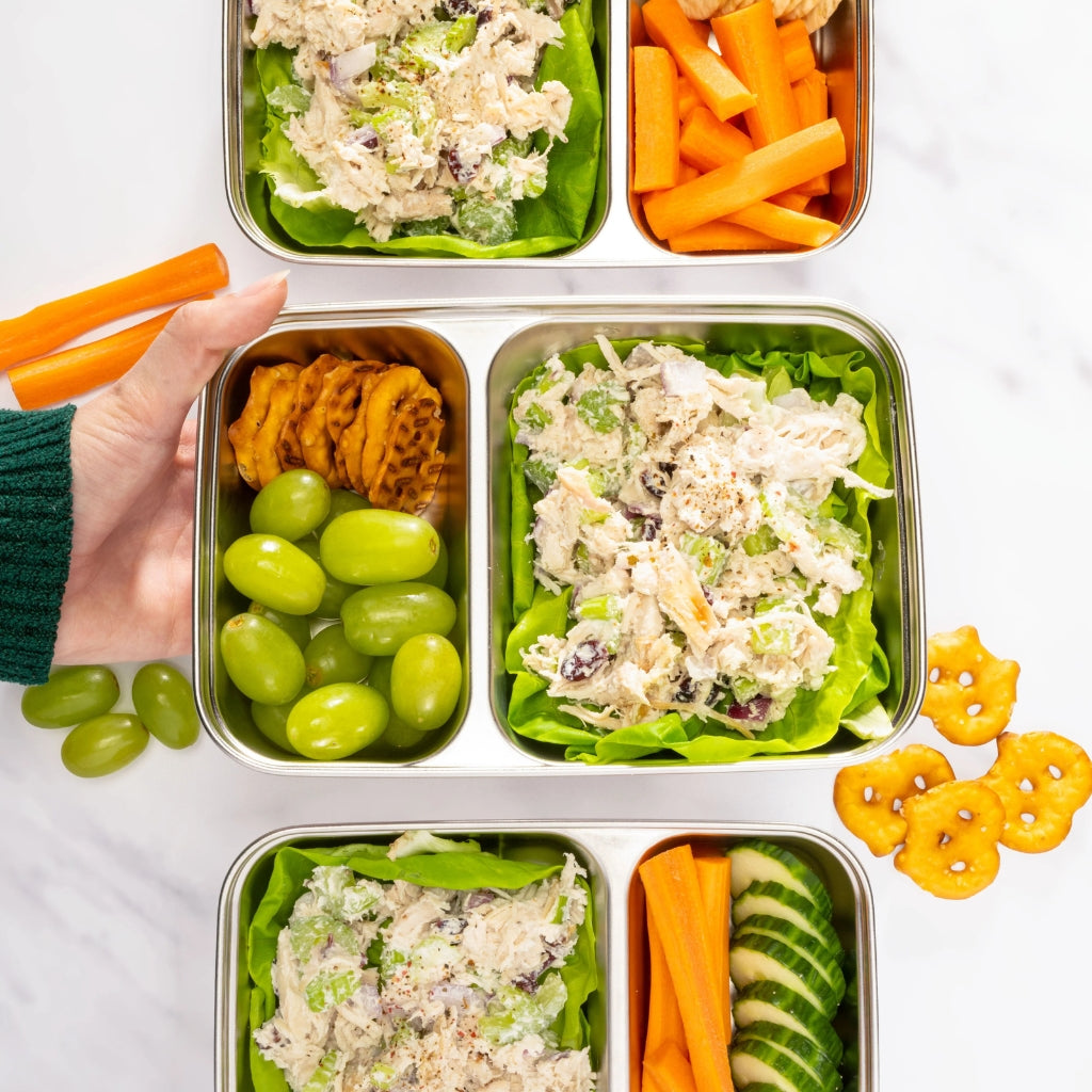 Three metal bento-style lunch boxes arranged on a white surface, filled with salad, carrot sticks, green grapes, waffles, and sliced cucumber. A hand wearing a dark green sleeve holds carrot sticks near the top left.