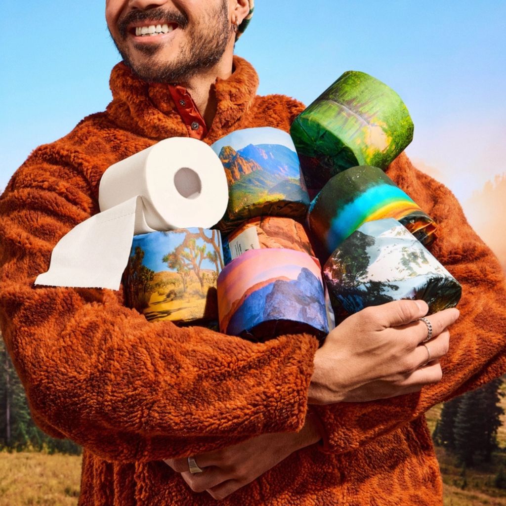 A man in a brown sherpa fleece jacket holds a stack of toilet paper rolls printed with colorful landscape scenes, including mountains, forests, and sunsets, against an outdoor sky background.