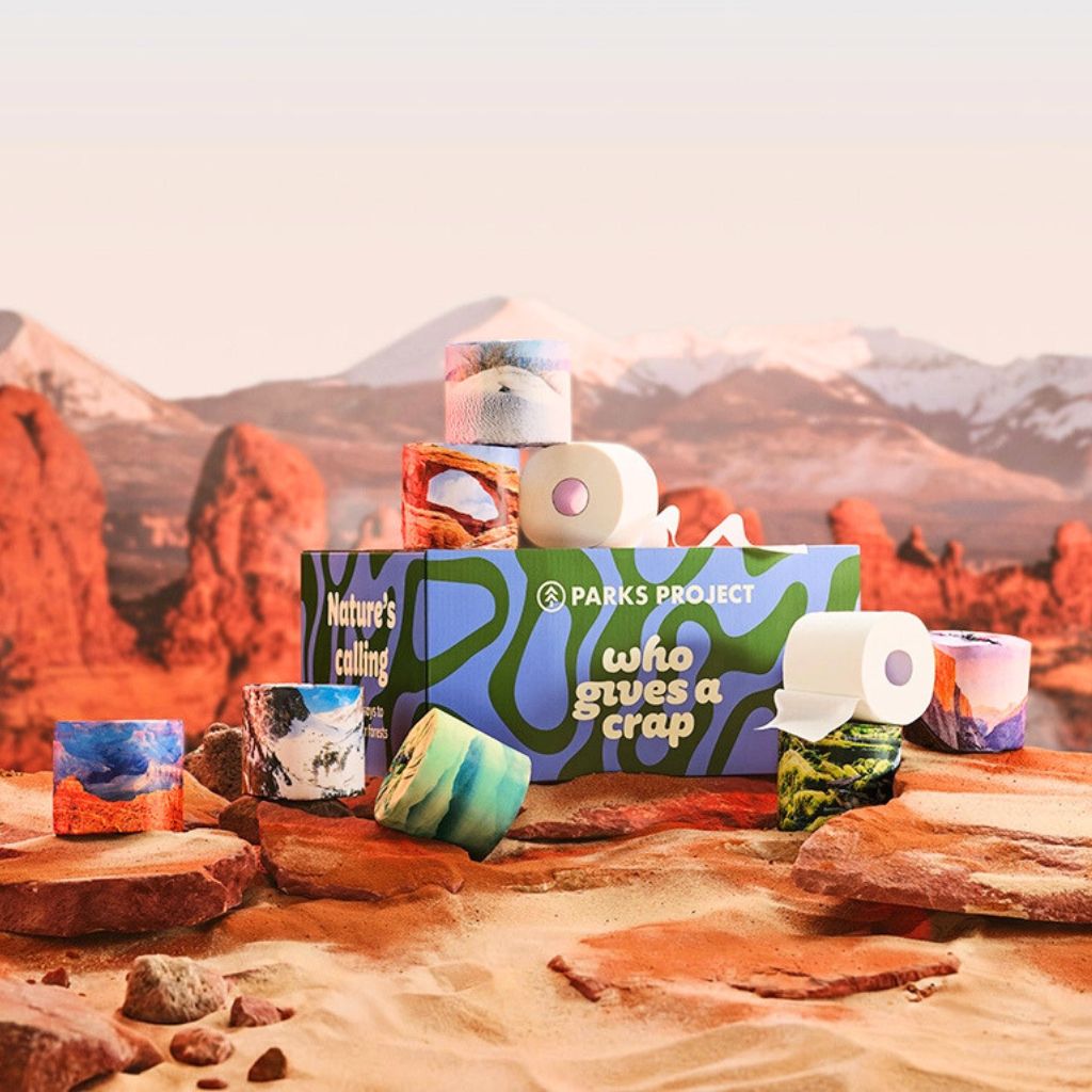 A Parks Project gift box surrounded by assorted toilet paper rolls and small marbled candles, styled on a sandy rock surface with red desert formations and mountains in the background.