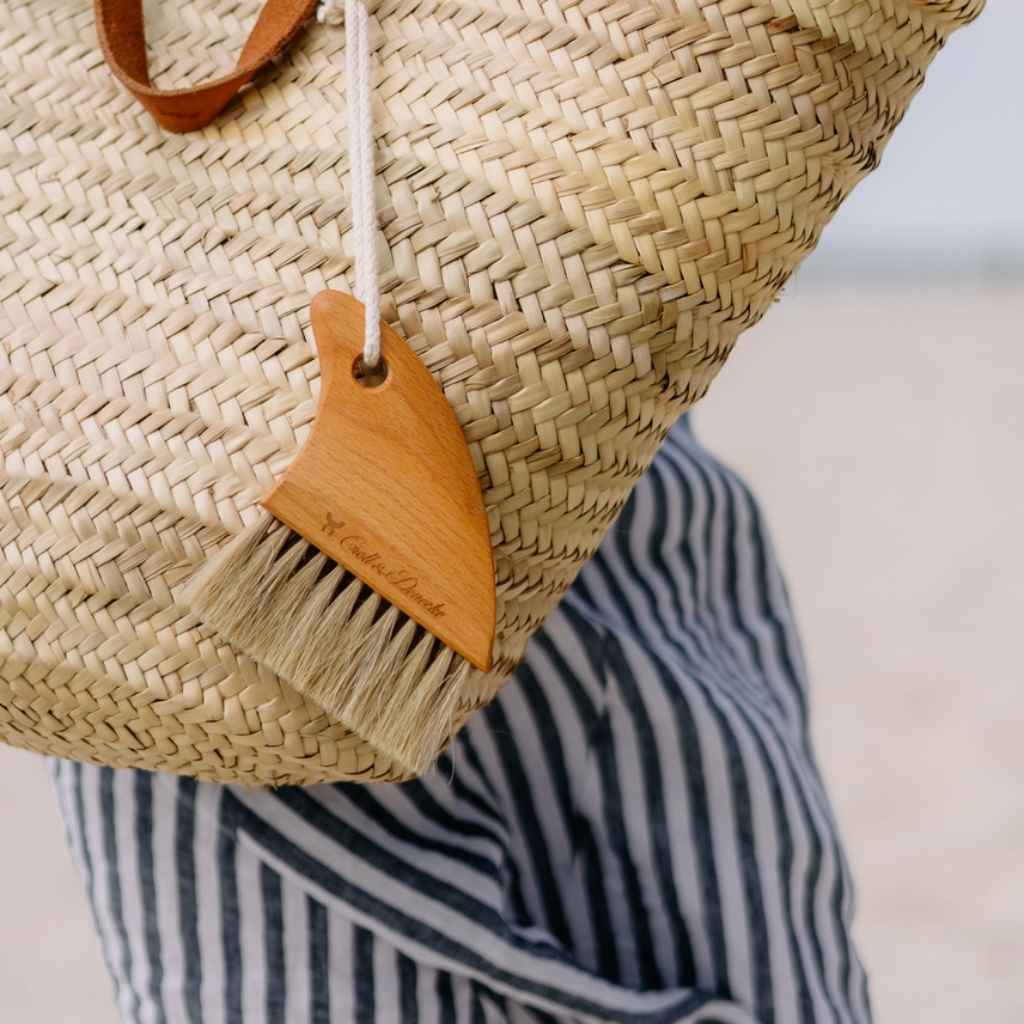 Sand brush for hands and feet - at the beach! Made by Croll &amp; Denecke out of beechwood. Carved into the shape of a shark fin. Shown attached to a beach bag with included carabiner
