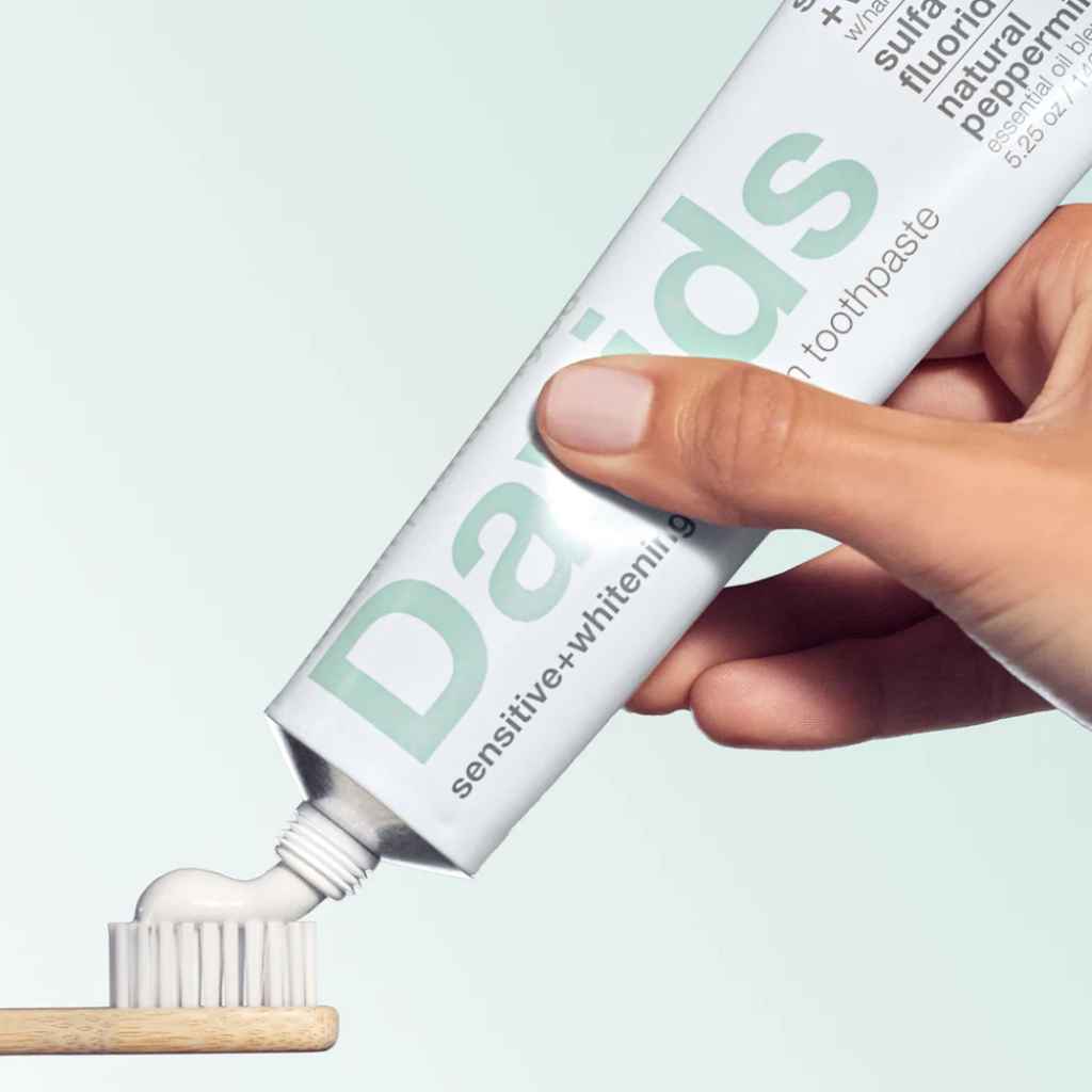 a hand squeezing a tube of Davids Premium Toothpaste 5.25oz in Sensitive + Whitening on to a bamboo toothbrush