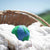 Single wool dryer ball designed to look like the earth in blue and green. Shown among clean laundry in basket, outside. Made by Friendsheep
