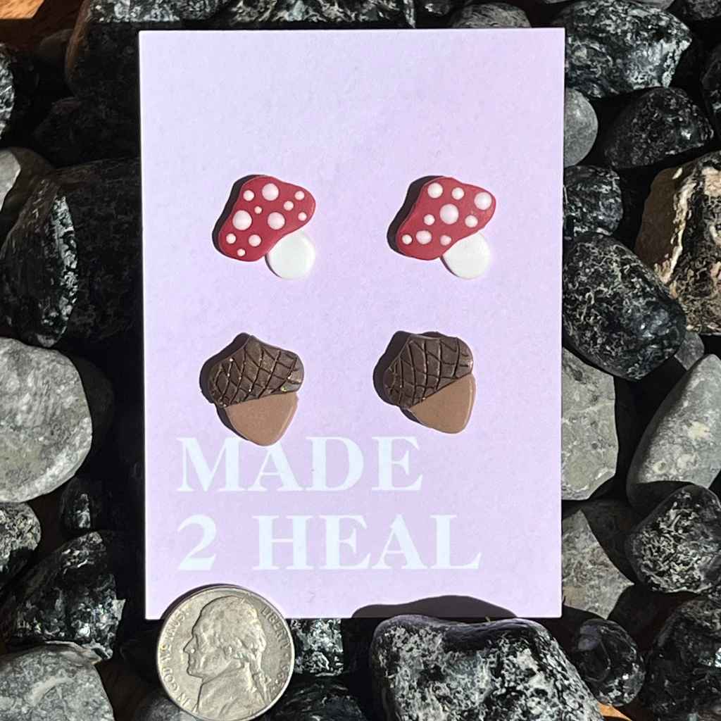 acorn and mushroom stud earrings on purple paper label on bed of rocks