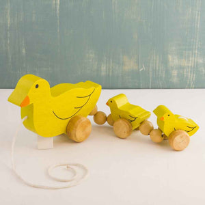 Wooden Pull-Along Toy by Mr. Ellie Pooh shown with three yellow ducks in a row. Made of sustainable Albizia wood from Sri Lanka, Fairtrade certified. US compliant coatings.