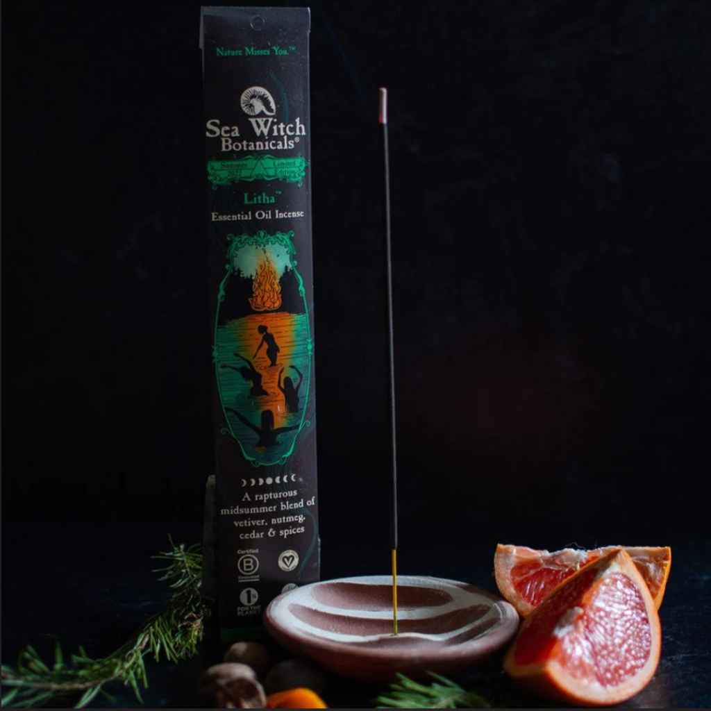 black vertical paper package with all-natural incense sticks made by Sea Witch Botanicals. Black package with green and orange design. Scent called Litha. 20 sticks. Shown next to one incense stick in a holder, with some fruit and greenery