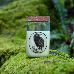 glass jar candle with cork lid, raven design on the front, pictured sitting on a mossy branch