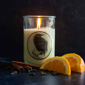 glass jar candle, lit, with black rave on the front, pictured with orange slices, cinnamon sticks, and cloves on a table