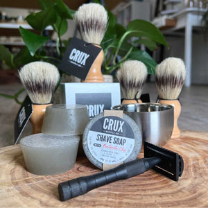 A collection of shaving supplies grouped together on a wood platter, includes black razor, shave soap, shave bowl, natural bristle shave brush.