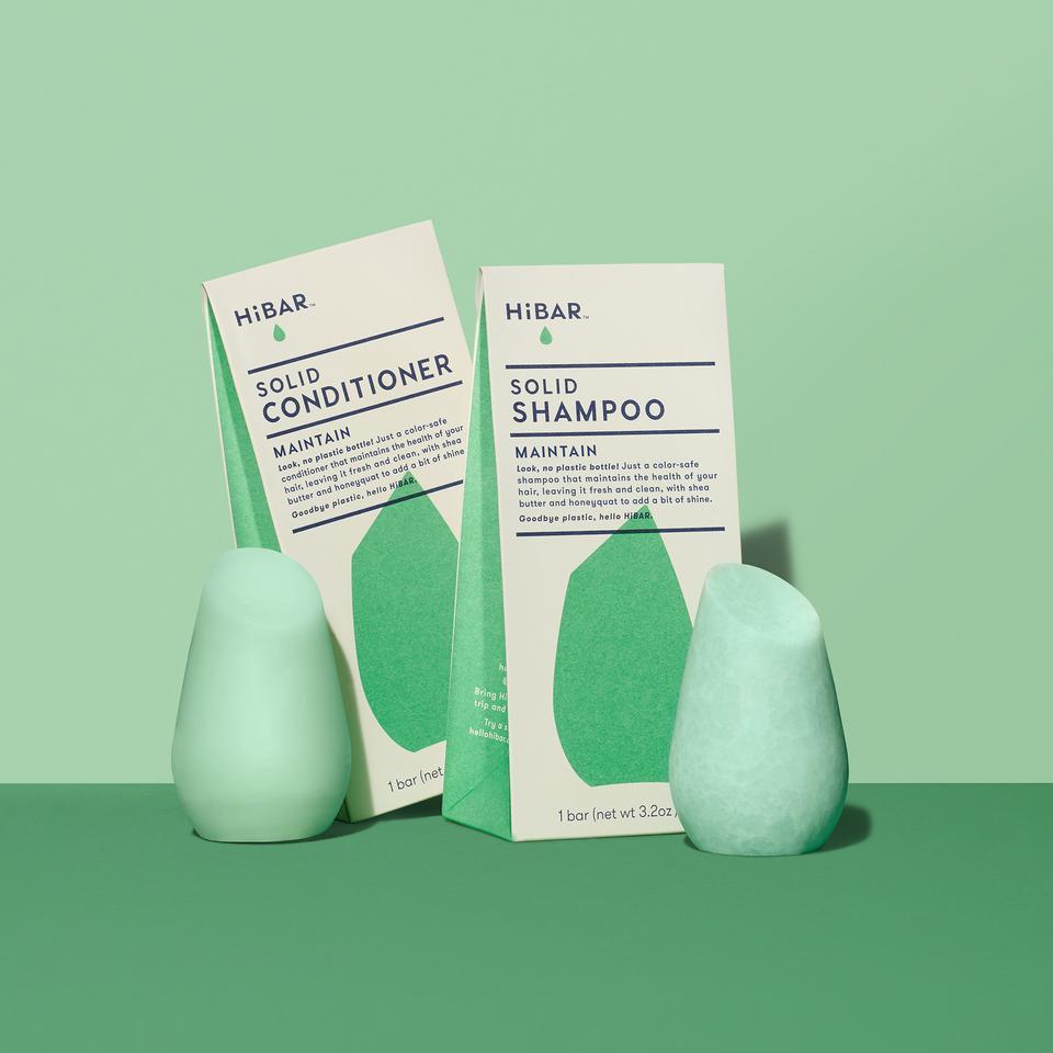 3.2 oz mint green solid conditioner bar, 3.2 oz mint green shampoo bar, displayed in front of respective packages on green back drop and dark green tabletop. shape of resembles and tapered, bulb like cylinder with flat, angled top