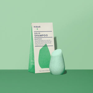  3.2 oz mint green shampoo bar, displayed in front of packaging on green back drop and dark green tabletop. shape of resembles and tapered, bulb like cylinder with flat, angled top