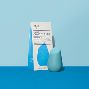 3.2 oz cyan conditioner bar, displayed in front of packaging on powder blue backdrop and dark blue tabletop. shape of resembles and tapered, bulb like cylinder with flat, angled top