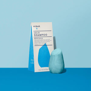 3.2 oz cyan shampoo bar, displayed in front of packaging on powder blue backdrop and dark blue tabletop. shape of resembles and tapered, bulb like cylinder with flat, angled top