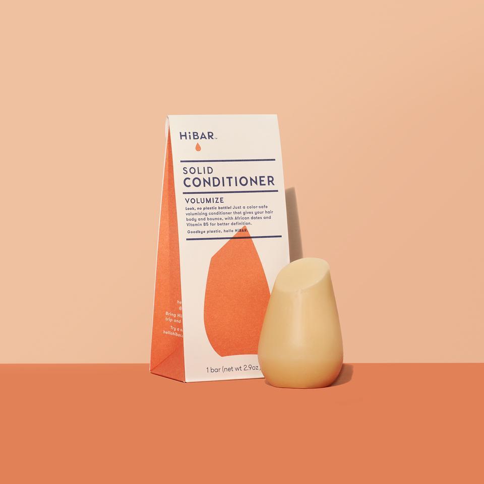 3.2 oz mango colored conditioner bar, displayed in front of packaging on soft orange backdrop and dark orange tabletop. shape of resembles and tapered, bulb like cylinder with flat, angled top