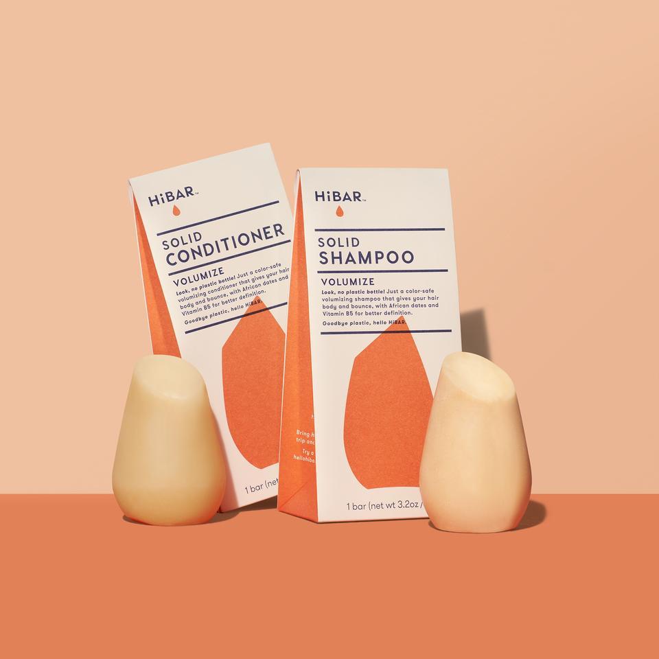 3.2 oz mango colored solid conditioner bar, 3.2 oz mango colored shampoo bar, displayed in front of respective packages on soft orange backdrop and dark orange tabletop. shape of resembles and tapered, bulb like cylinder with flat, angled top