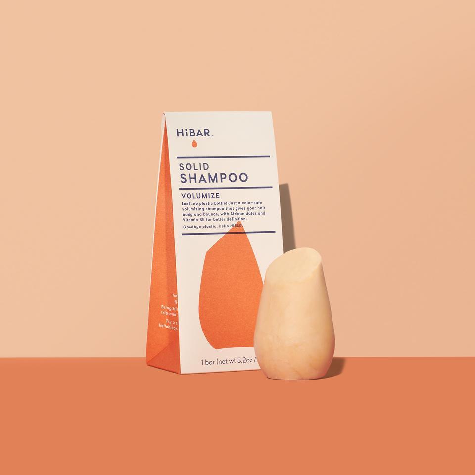3.2 oz mango colored shampoo bar, displayed in front of packaging on soft orange backdrop and dark orange tabletop. shape of resembles and tapered, bulb like cylinder with flat, angled top