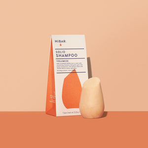 3.2 oz mango colored shampoo bar, displayed in front of packaging on soft orange backdrop and dark orange tabletop. shape of resembles and tapered, bulb like cylinder with flat, angled top