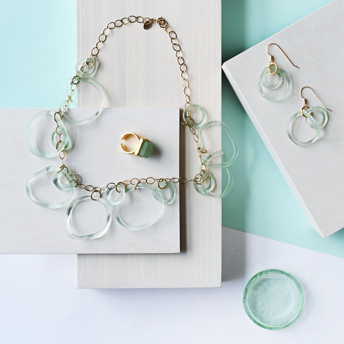 Gold band cocktail ring with square green stone next to a necklace and earring made from recycled glass
