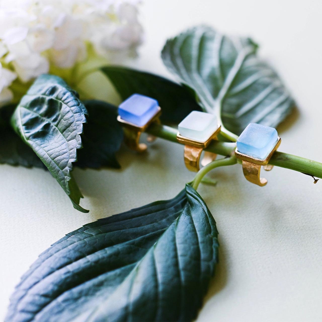 Three gold band rings with square blue stones sitting on the stem of a cut hydrangea