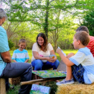 two adults and three children sit outdoors on hay bails, playing wildcraft game, happy