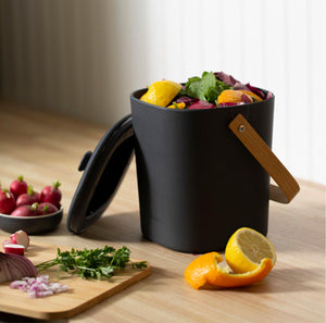 orange and lemon peels, baby radishes and a cutting board with cilantro and diced onion surrounding a black compost bin with a wooden handle. the bin is filled to the brim with fresh fruit and vegetable left overs, ready to be transported to compost pile 