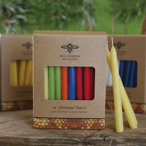 cardboard pack of 45 multicolored hanukkah tapered beeswax candles, dispalyed in front of two other packages on a wooden tabletop with a green forest backdrop