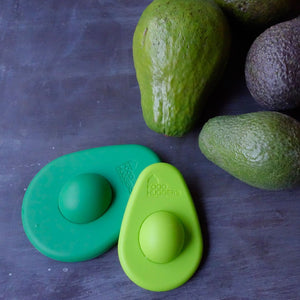 2 reusable green silicone avocado savers, one small and light green, one large and dark green, next to 3 ripe avacodos