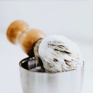 natural bristle shave brush lathered up with shave soap resting on stainless steel shave bowl against a white background.
